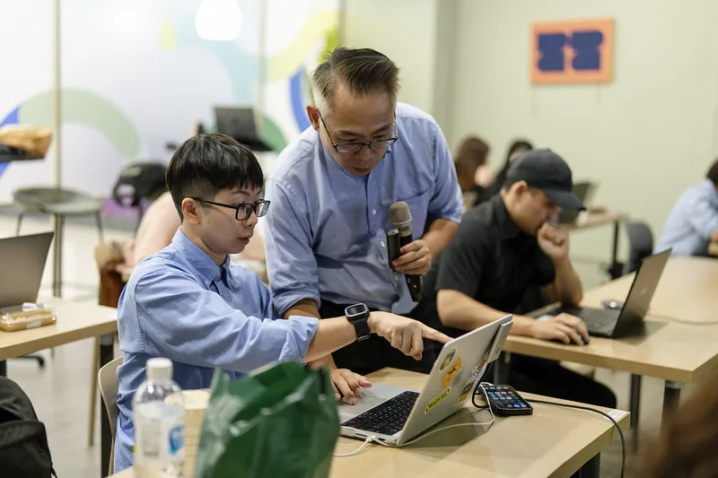 Yu Wen-Hao leaning in beside a participant who is pointing at his own laptop screen