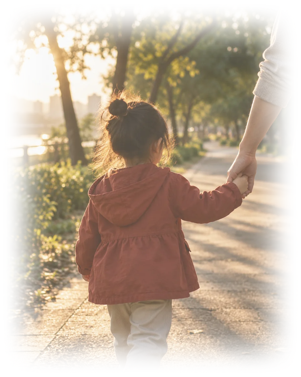 A small child in a brick-red jacket, seen from behind, walking along a park path at dusk holding an adult's hand.